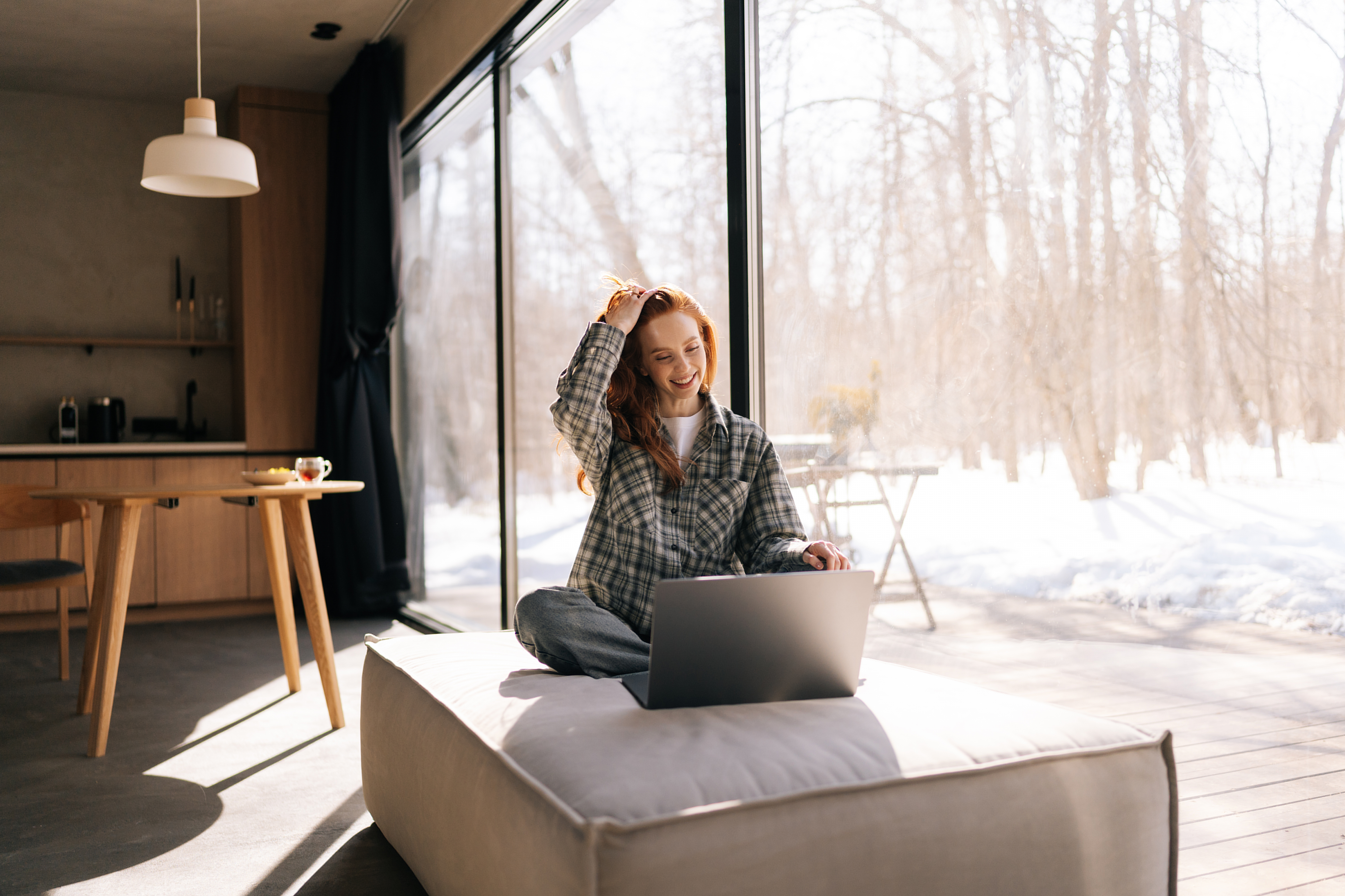 Woman sitting on ottoman using laptop by large window